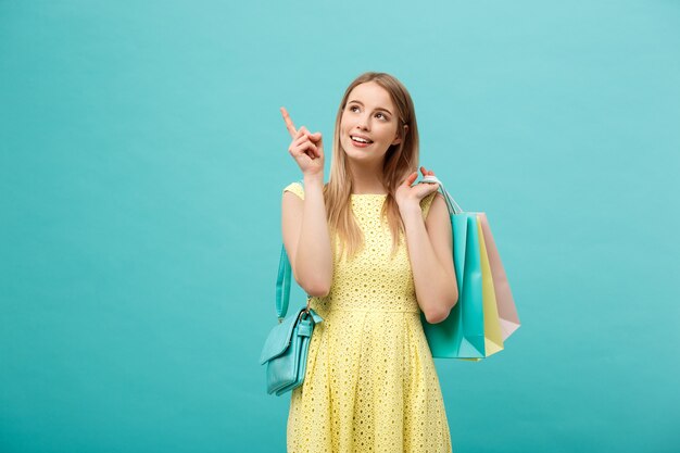 Hermosa joven con bolso de compras en vestido amarillo apunta a algo con su dedo.