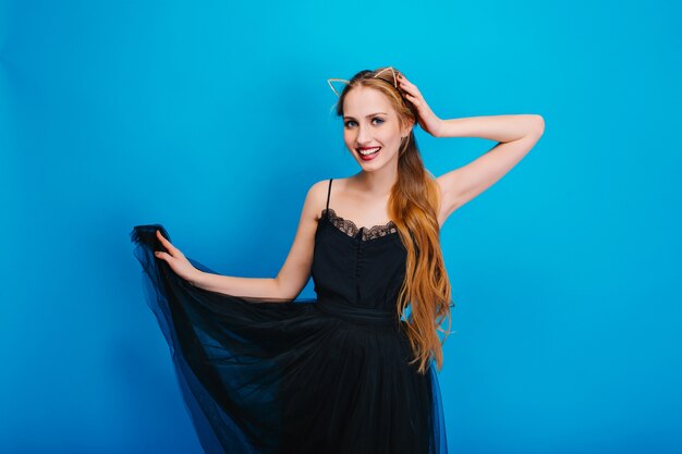 Hermosa joven en bonito vestido negro esponjoso posando en la fiesta, sonriendo. Tiene el pelo largo y ondulado, y lleva diadema de orejas de gato con cristales.