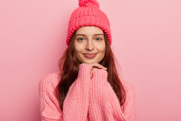 Hermosa joven de cabello oscuro mantiene las manos debajo de la barbilla, viste ropa abrigada de invierno, mira con alegría a la cámara, tiene una belleza natural sin maquillaje.