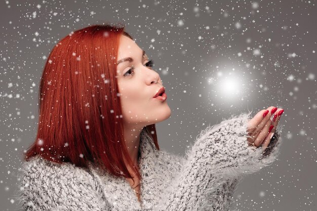 Hermosa joven con cabello rojo y uñas rojas sosteniendo sus manos juntas y soplando una bola de luz blanca Mujer bonita en suéter gris atrapando estrellas y pidiendo deseos cuando nieva