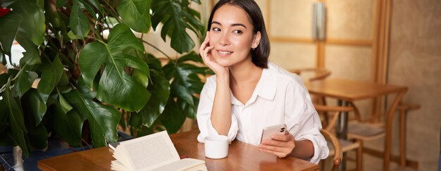 Foto gratuita hermosa joven en la cafetería con una taza de café y un libro sosteniendo un teléfono inteligente usando un teléfono móvil