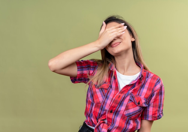 Una hermosa joven con una camisa a cuadros que cubre los ojos con la mano sobre una pared verde