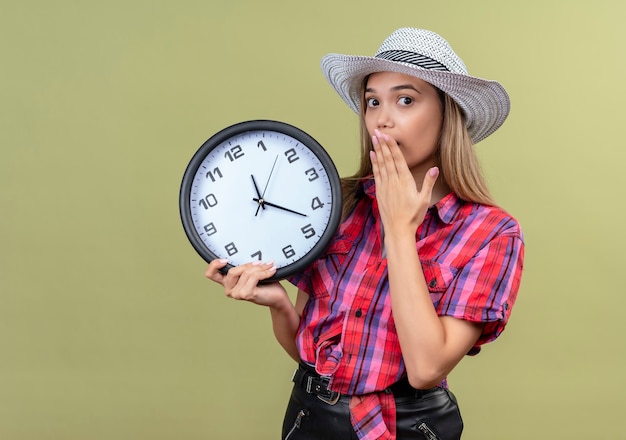 Una hermosa joven en una camisa a cuadros con sombrero sosteniendo el reloj de pared mientras mantiene la mano en la boca en una pared verde