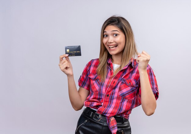 Una hermosa joven en camisa a cuadros sonriendo mientras muestra la tarjeta de crédito con el puño cerrado en una pared blanca