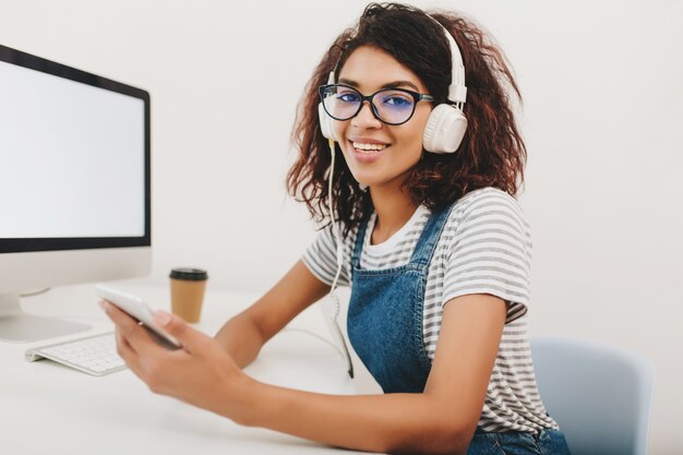 Hermosa joven en camisa a rayas sentada en la oficina y sosteniendo el teléfono inteligente en la mano esperando la llamada