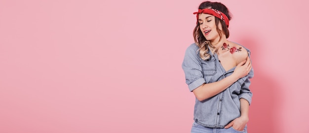 Hermosa joven en camisa vaquera con calcomanía agua tatuaje flores pegatina y sonriente sobre fondo rosa