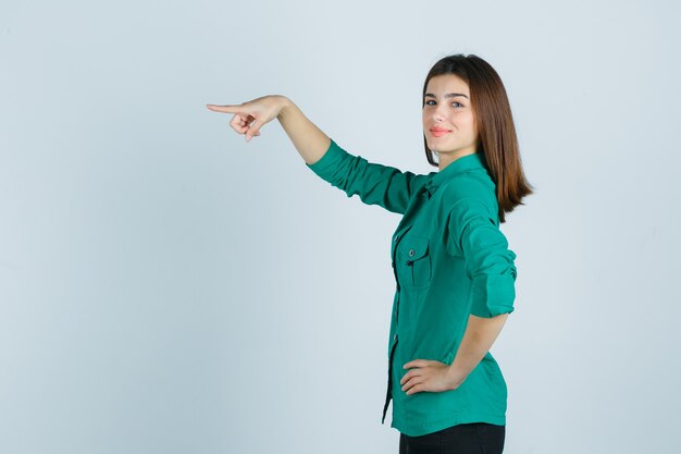 Hermosa joven en camisa verde apuntando hacia adelante y mirando alegre.