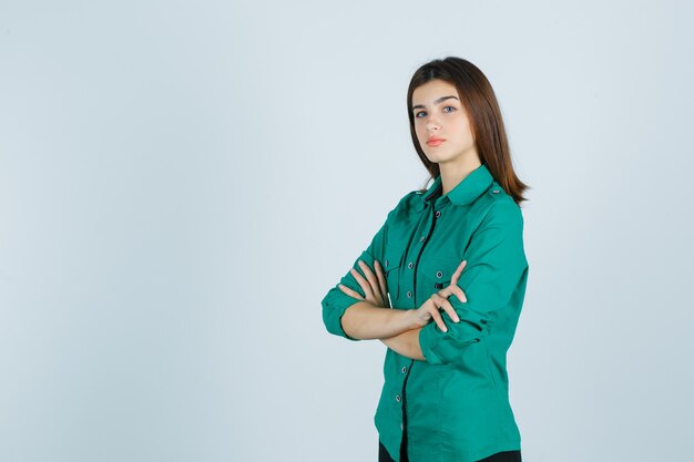 Hermosa joven en camisa verde sosteniendo los brazos cruzados y mirando seria, vista frontal.