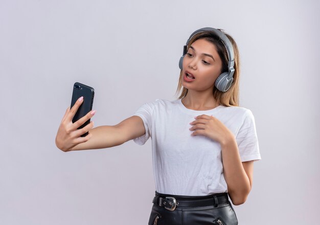 Una hermosa joven en camiseta blanca usando audífonos tomando un selfie con smartphone en una pared blanca