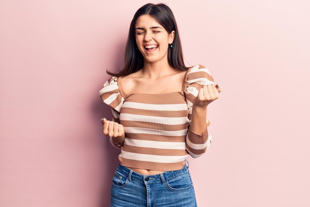 Hermosa joven con una camiseta casual a rayas celebrando sorprendida y asombrada por el éxito con los brazos levantados y los ojos cerrados