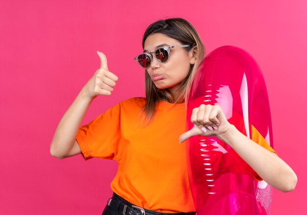 Una hermosa joven con una camiseta naranja con gafas de sol mostrando los pulgares hacia arriba y hacia abajo mientras sostiene el anillo inflable rosa en una pared rosa