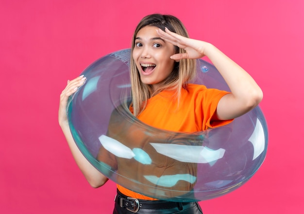 Foto gratuita una hermosa joven en una camiseta naranja mirando mientras está de pie sobre un anillo inflable en una pared rosa