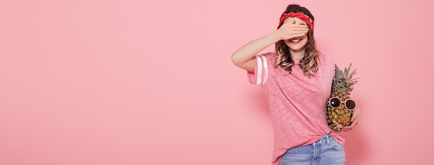 Hermosa joven en camiseta rosa, cubre su rostro, sostiene piña