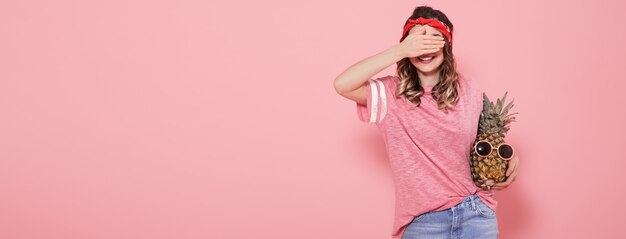 Hermosa joven en camiseta rosa, cubre su rostro, sostiene piña