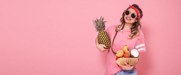 Hermosa joven en camiseta rosa y gafas, sostiene una bolsa de paja llena de frutas sobre fondo rosa