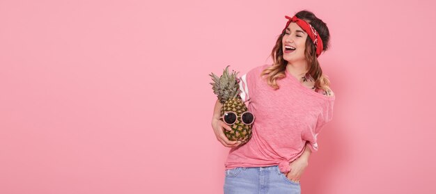 Hermosa joven en camiseta rosa, sonriendo con piña en pared rosa