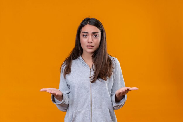 Hermosa joven con capucha gris en emoción desesperada de pie sobre fondo naranja