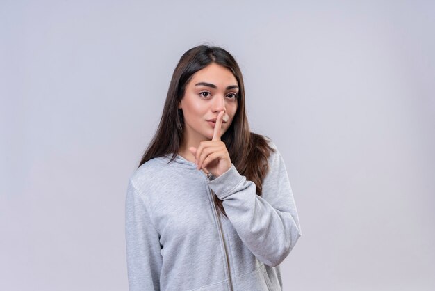 Hermosa joven con capucha gris haciendo gesto de silencio de pie sobre fondo blanco.