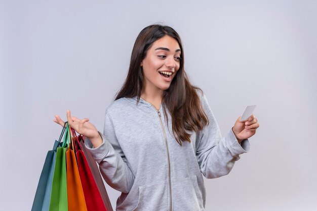 Foto gratuita hermosa joven con capucha gris mirando al papel con una sonrisa en la cara sosteniendo paquetes de pie sobre fondo blanco.