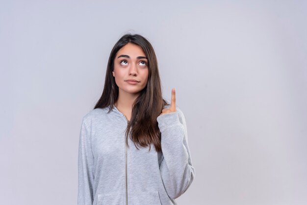 Hermosa joven con capucha gris mirando hacia arriba y apuntando hacia arriba de pie sobre fondo blanco.