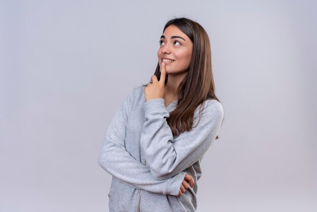 Hermosa joven con capucha gris mirando hacia arriba con el dedo en la barbilla sonrisa en la cara de pie sobre fondo blanco.