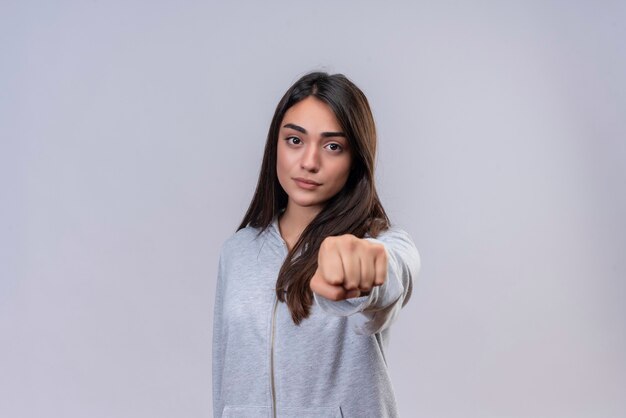 Hermosa joven con capucha gris mirando a la cámara apretando el puño a la cámara con expresión enojada de pie sobre fondo blanco.