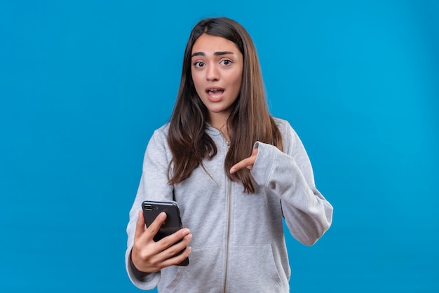 Hermosa joven con capucha gris mirando a la cámara con cara de sorpresa y apuntando al teléfono de pie sobre fondo azul.