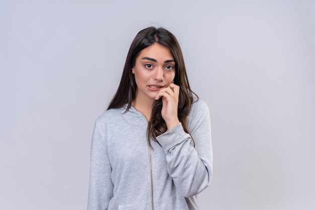 Hermosa joven con capucha gris mirando a la cámara con confundido en la cara tocando la mejilla de pie sobre fondo blanco.