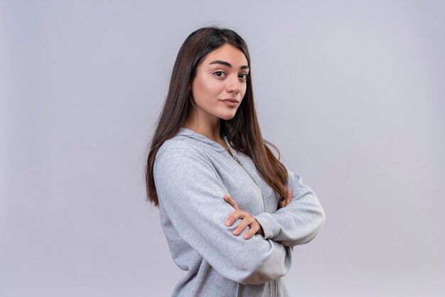 Hermosa joven con capucha gris mirando a la cámara con emoción agradable cruzando las manos de pie sobre fondo blanco.