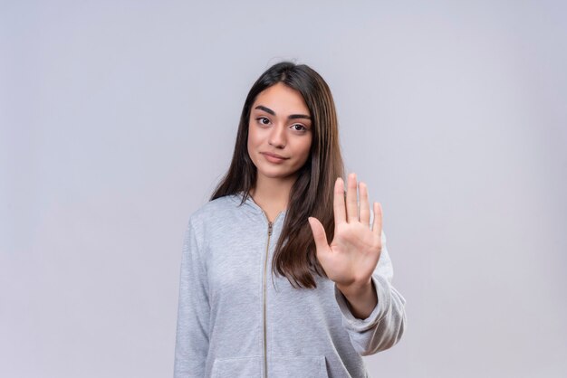 Hermosa joven con capucha gris mirando a cámara con expresión desagradable haciendo gesto de parada de pie sobre fondo blanco.