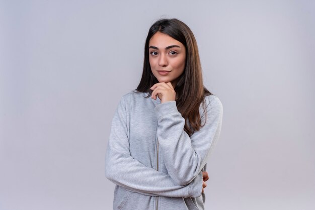 Hermosa joven con capucha gris mirando a la cámara con expresión de placer con la mano en la barbilla de pie sobre fondo blanco.