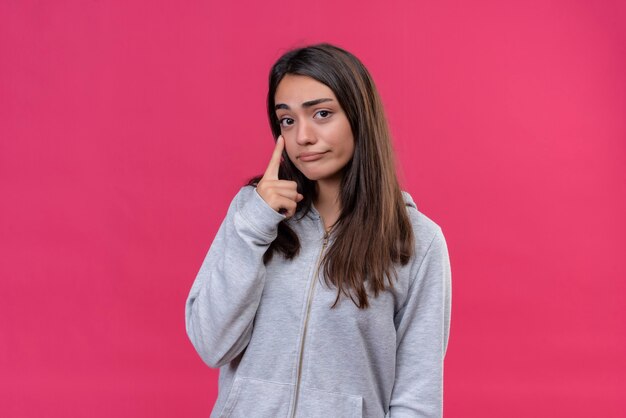 Hermosa joven con capucha gris mirando a la cámara con expresión triste tocando la mejilla de pie sobre fondo rosa