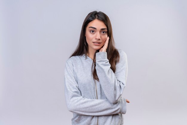 Hermosa joven con capucha gris mirando a la cámara inquieta en la cara tocando la mejilla de pie sobre fondo blanco.