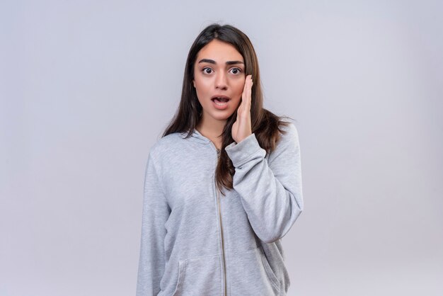 Hermosa joven con capucha gris mirando a la cámara con la mano de cara conmocionada cerca de la boca de pie sobre fondo blanco.