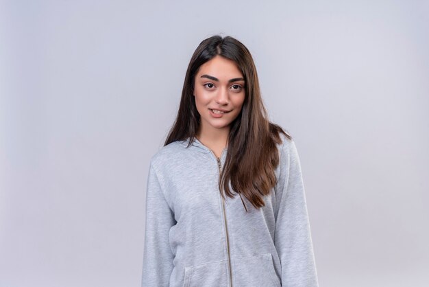 Hermosa joven con capucha gris mirando a la cámara mordiendo el labio de pie sobre fondo blanco.