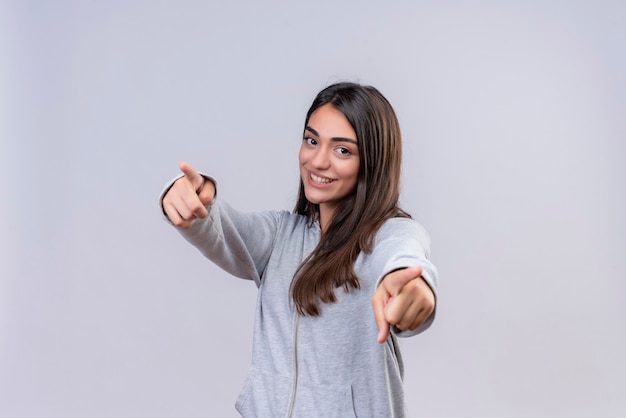 Hermosa joven con capucha gris mirando a la cámara con una sonrisa en la cara alegre y apuntando a la cámara de pie sobre fondo blanco.