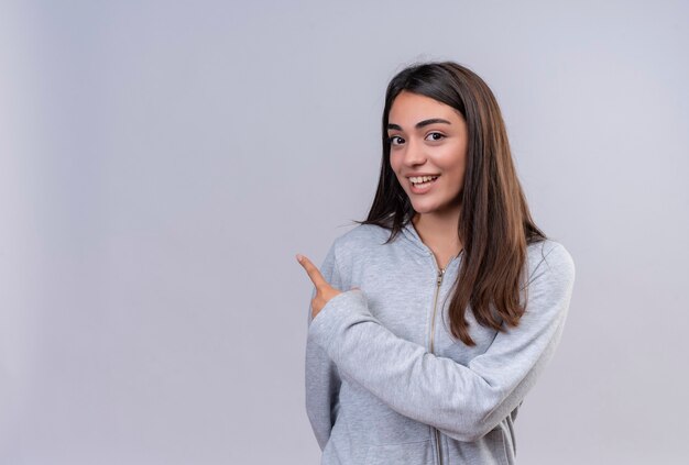 Hermosa joven con capucha gris mirando a la cámara con una sonrisa en la cara apuntando hacia atrás de pie sobre fondo blanco.