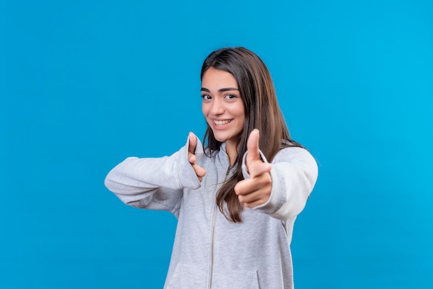 Hermosa joven con capucha gris mirando a la cámara con una sonrisa en la cara apuntando a la cámara de pie sobre fondo azul.