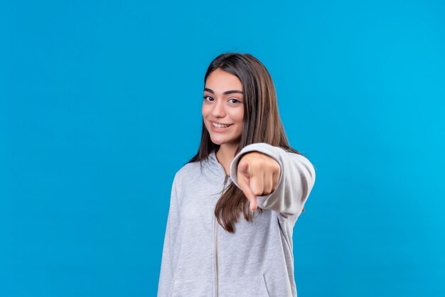 Hermosa joven con capucha gris mirando a la cámara con una sonrisa en la cara apuntando a la cámara de pie sobre fondo azul.