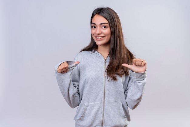 Hermosa joven con capucha gris mirando a la cámara con una sonrisa en la cara apuntando a ti mismo de pie sobre fondo blanco.