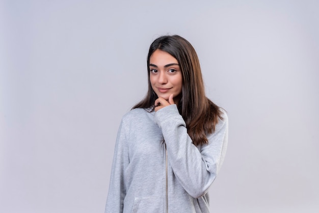 Hermosa joven con capucha gris mirando a la cámara sonrisa en la cara con el dedo en la barbilla de pie sobre fondo blanco.