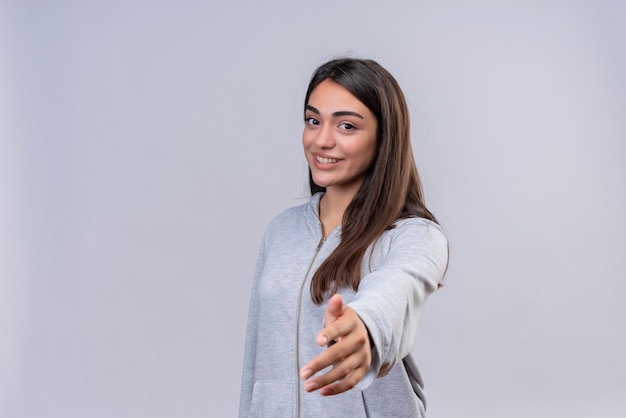 Hermosa joven con capucha gris mirando a la cámara con una sonrisa en la cara extendiendo su mano de pie sobre fondo blanco.