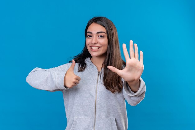 Hermosa joven con capucha gris mirando a la cámara con una sonrisa en la cara haciendo como y pare el gesto de pie sobre fondo azul.