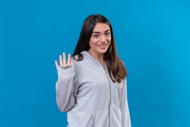 Hermosa joven con capucha gris mirando a la cámara con una sonrisa en la cara y haciendo gesto de hola de pie sobre fondo azul.