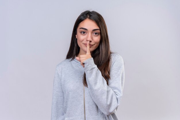 Hermosa joven con capucha gris mirando a la cámara con una sonrisa en la cara haciendo gesto de silencio de pie sobre fondo blanco.