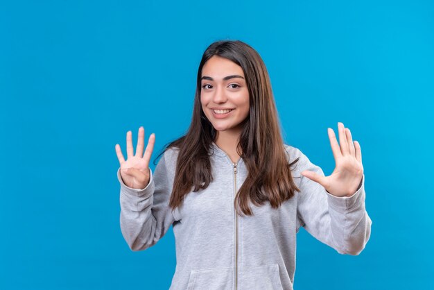 Hermosa joven con capucha gris mirando a la cámara con una sonrisa en la cara mostrando cinco y cuatro números en las manos de pie sobre fondo azul.