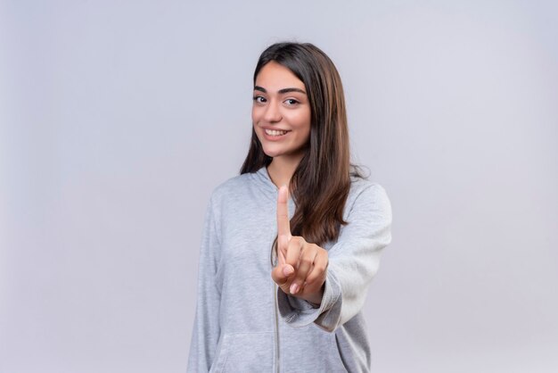 Hermosa joven con capucha gris mirando a la cámara sonrisa en la cara mostrando un número de pie sobre fondo blanco.