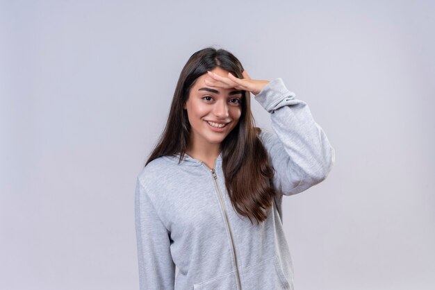 Hermosa joven con capucha gris mirando a la cámara con una sonrisa en la cara poniendo la mano en la frente de pie sobre fondo blanco.