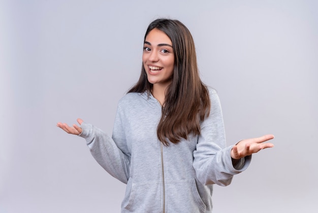 Hermosa joven con capucha gris mirando a la cámara con una sonrisa en la cara sin respuesta, extendiendo las palmas sobre fondo blanco.