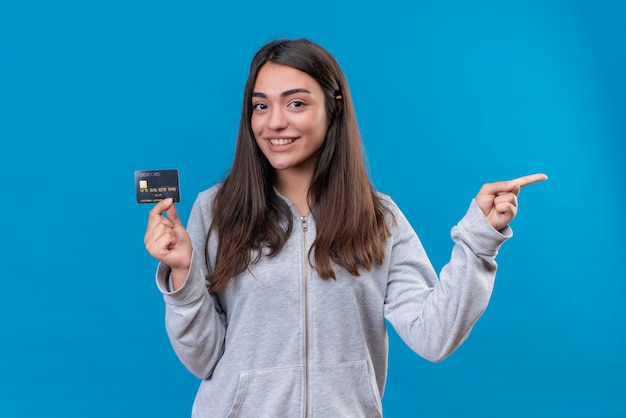 Hermosa joven con capucha gris mirando a la cámara con una sonrisa en la cara sosteniendo la tarjeta de crédito y apuntando con otra mano sobre fondo azul.